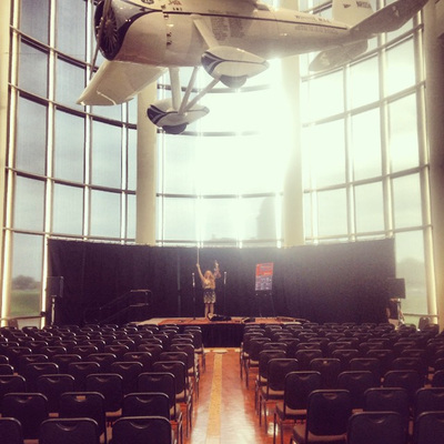 Minna Biggs stands on stage preshow at the Oklahoma History Museum. Wiley Post’s famous Winnie Mae airplane hangs overhead.