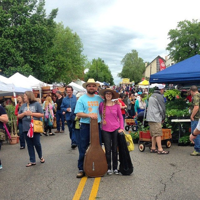 Casey & Minna with fiddle and guitar in cases at the Sand Springs Herbal Affair. Colorful tents and people fill the street.