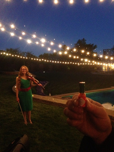 At twilight, elegantly dressed in green, Minna Biggs holds a violin, standing on lush grass, pool side at a private party in Nichols Hills. In the foreground a hand holds a mini Tabasco sauce bottle. String lights twinkle overhead.