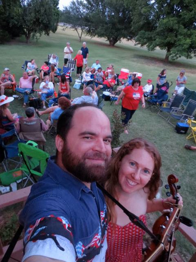 Oklahoma fiddle and guitar duo, Casey & Minna, take a selfie at the 2014 Deer Creek 4th of July private party. A hometown crowd dressed in red, white, and blue mingle among lawn chairs on the grass. 